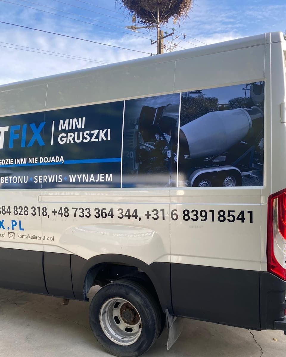 Lewy bok srebrnego busa od tyłu: ciemny panel z napisem MINI GRUSZKI i zdjęciami, trzy numery telefonów, tylne koło, nad dachem bocianie gniazdo.